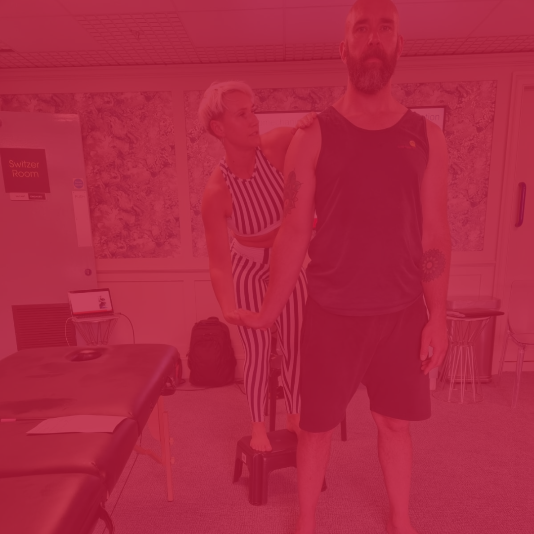 Man and woman in a class learnering about biomechanics. Man is staning, woman is behind him, he has his arm out straight and she is looking at his arm whilst holding it.