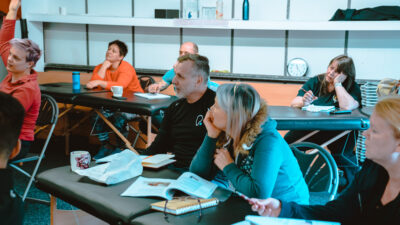 Men nd women in a classroom setting, all wearing gym gear, and listening intently with pens and paper in front of them.