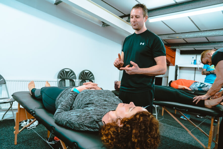 man counting on this fingers, wearing a black t-short, standing over a woman on a massage table who has her eyes closed and arms folded.