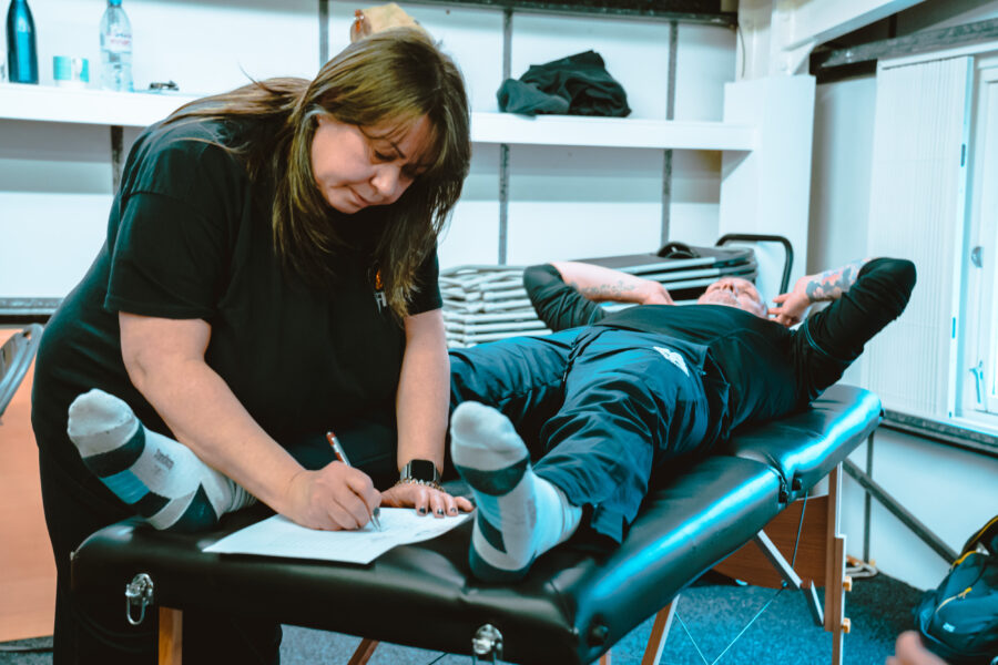 woman working on notes whislt assessing a client who is a man, laying on a massage table.