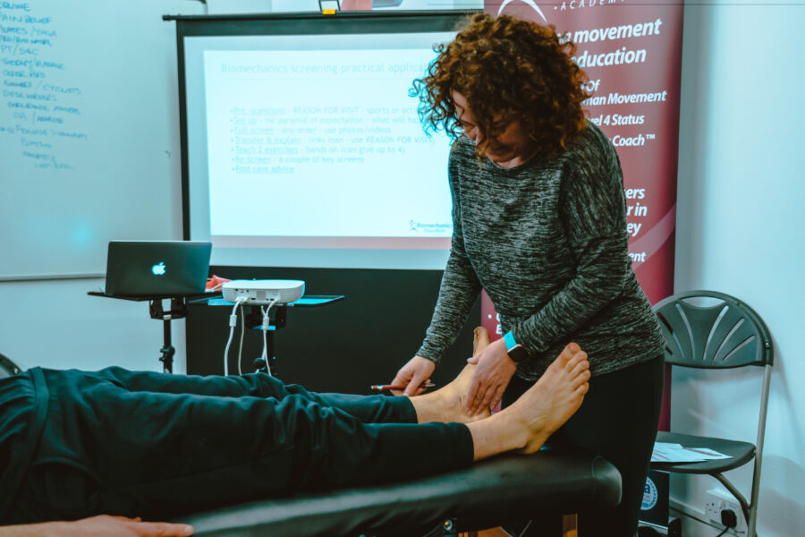 Woman with red curly hair, wearing a long sleeve grey top, and black leggings, treating a persons post posture as they are laying on a massage bench.