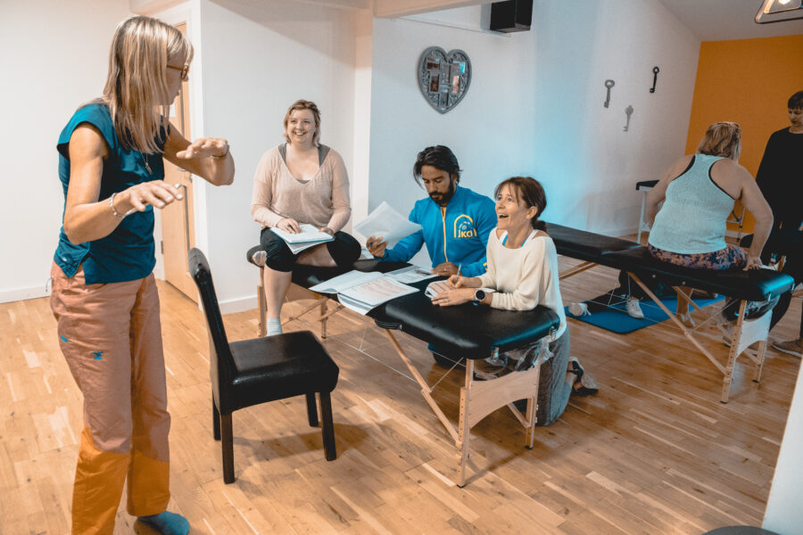 Lenrs of intrinisc biomechanics. one sat on a massage table, two dat in chairs leaning on the table with their arms and taking notes. There is an animated woman explainging something to them, she is standing up in front of them and they are smiling at her.