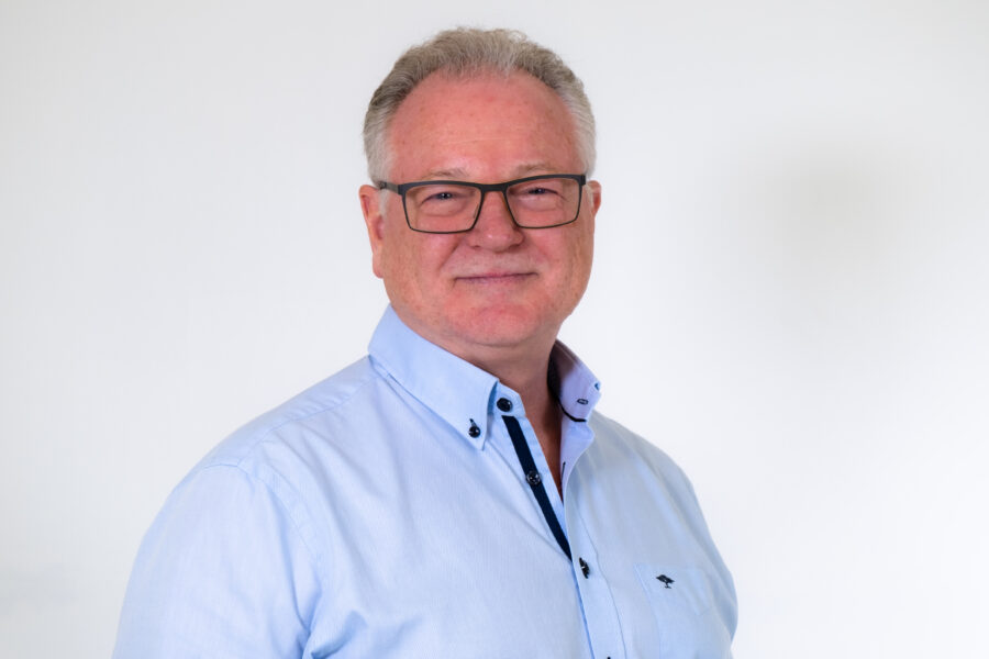 Martin Haines, wearing a light blue shirt, smilng at the camera, white/grey hair, black rimmed glasses, white older male.